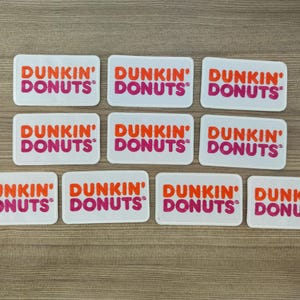 Op de afbeelding: Negen witte rechthoekige stickers met de tekst "DUNKIN' DONUTS" in oranje en roze letters.