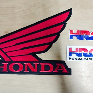 May include: Embroidered Honda wing logo patch in red and black, with two smaller HRC patches. One HRC patch has the text "HONDA RACING". The patches are on a light wood surface.