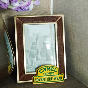 May include: A brown picture frame with a photo of the Space Needle in Seattle, Washington. The frame has a gold trim and a brown leather-like texture. Below the frame is a yellow and green patch that reads "Camel Trophy Adventure Wear."