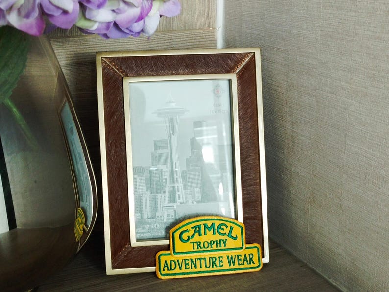 May include: A brown picture frame with a photo of the Space Needle in Seattle, Washington. The frame has a gold trim and a brown leather-like texture. Below the frame is a yellow and green patch that reads "Camel Trophy Adventure Wear."