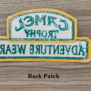 May include: A white patch with yellow and green trim. The text on the patch reads "Camel Trophy Adventure Wear".