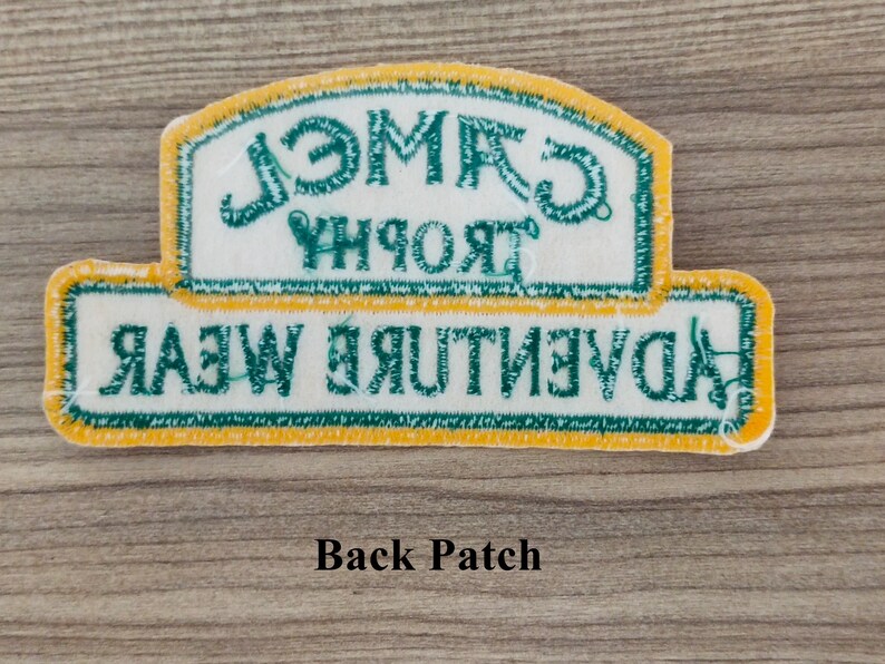 May include: A white patch with yellow and green trim. The text on the patch reads "Camel Trophy Adventure Wear".