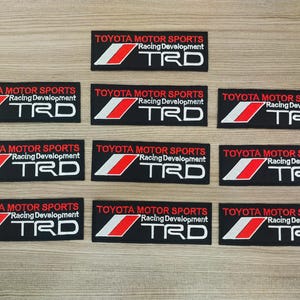 Könnte beinhalten: Neun schwarze und weiße Patches mit roten Akzenten. Jeder Patch hat den Text "TOYOTA MOTOR SPORTS" und "Racing Development" mit dem Logo "TRD" in Weiß.