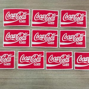 Può includere: Dieci toppe rettangolari rosse con loghi Coca-Cola bianchi e la parola "Coke" stampata sotto ogni logo.