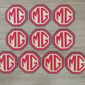 Op de afbeelding: Acht achtkantige rode en witte patches met de letters "MG" in wit op een rode achtergrond.