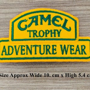 May include: Yellow and green embroidered patch with the text "Camel Trophy Adventure Wear". The patch is approximately 10 cm wide and 5.4 cm high.