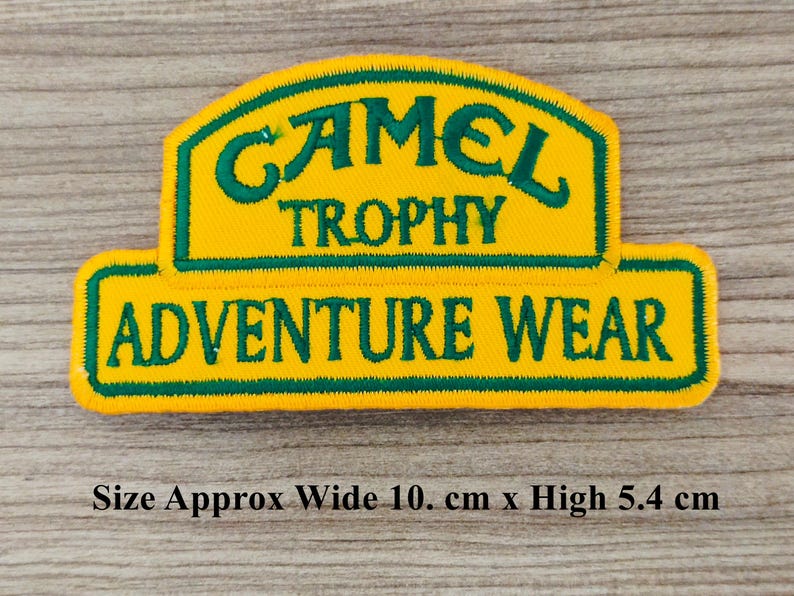 May include: Yellow and green embroidered patch with the text "Camel Trophy Adventure Wear". The patch is approximately 10 cm wide and 5.4 cm high.