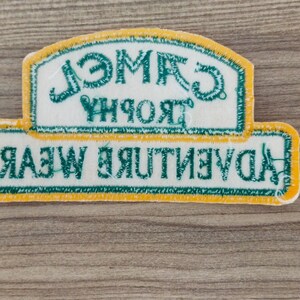 May include: A white patch with green and yellow trim. The patch has the text "Adventure Wear" in green and "Trophy Camel" in green, reversed.