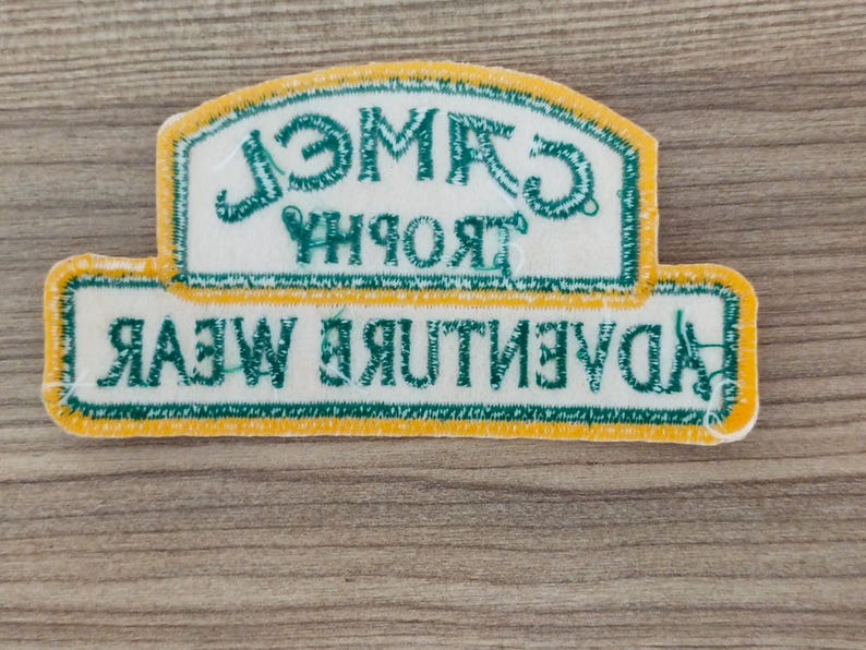 May include: A white patch with green and yellow trim. The patch has the text "Adventure Wear" in green and "Trophy Camel" in green, reversed.