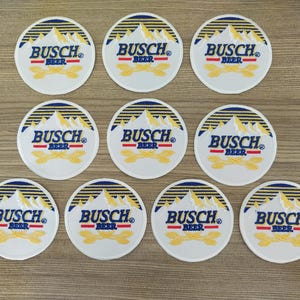 Puede incluir: Nueve parches redondos blancos con bordados azules y dorados. Cada parche presenta el texto "Busch Beer" con un diseño de cordillera.