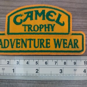 May include: A yellow and green embroidered patch with the text "CAMEL TROPHY ADVENTURE WEAR".