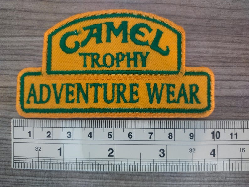 May include: A yellow and green embroidered patch with the text "CAMEL TROPHY ADVENTURE WEAR".