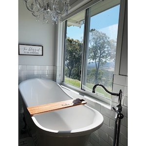 Op de afbeelding: Een witte vrijstaande badkuip met een houten badplank met de tekst "take a soak". De badkamer heeft witte metro tegels, een raam met uitzicht op bomen en een bord met de tekst "Too blessed to be stressed".