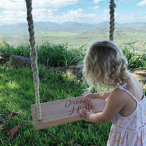 Op de afbeelding: Een houten schommel met de woorden "Dream High" erin gegraveerd hangt aan een dik touw. Een jong kind in een bloemenjurk raakt de schommel aan. De achtergrond toont een groen landschap en bergen onder een blauwe lucht.