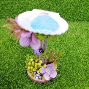 Op de afbeelding: Een miniatuur feeentuin sc&egrave;ne met een witte schelp met een blauw centrum, een houten basis en paarse bloemen.