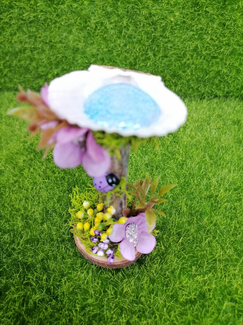 Op de afbeelding: Een miniatuur feeentuin sc&egrave;ne met een witte schelp met een blauw centrum, een houten basis en paarse bloemen.