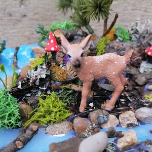 May include: A miniature diorama featuring a small fawn with white spots, standing near a miniature stream. The scene includes miniature trees, rocks, moss, and red and white spotted mushrooms, creating a whimsical forest setting.