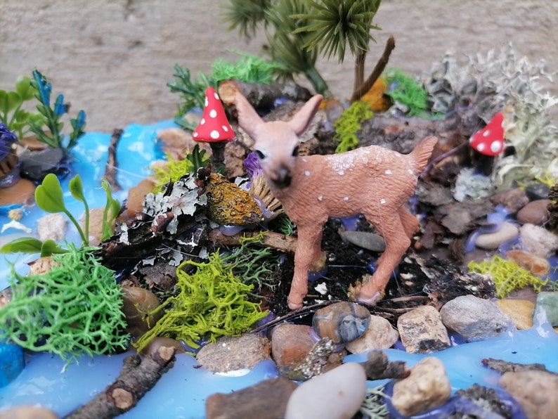 May include: A miniature diorama featuring a small fawn with white spots, standing near a miniature stream. The scene includes miniature trees, rocks, moss, and red and white spotted mushrooms, creating a whimsical forest setting.