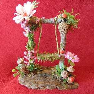 May include: A miniature wooden swing set with a moss-covered base and pink flowers. The swing set is decorated with ladybugs and pine cones.