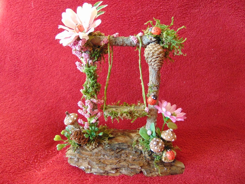 May include: A miniature wooden swing set with a moss-covered base and pink flowers. The swing set is decorated with ladybugs and pine cones.
