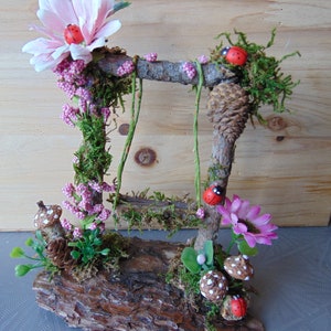 May include: A miniature fairy garden swing set made from twigs and moss, with pink flowers, green leaves, and red ladybugs. The swing set is attached to a wooden log base.