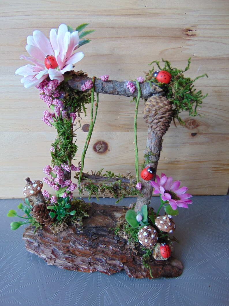 May include: A miniature fairy garden swing set made from twigs and moss, with pink flowers, green leaves, and red ladybugs. The swing set is attached to a wooden log base.
