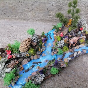 May include: A miniature woodland diorama featuring a flowing blue river, surrounded by green moss, rocks, and miniature trees. Red and white spotted mushrooms and a small deer figurine add detail to this nature-themed art piece.