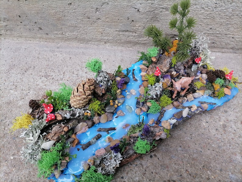 May include: A miniature woodland diorama featuring a flowing blue river, surrounded by green moss, rocks, and miniature trees. Red and white spotted mushrooms and a small deer figurine add detail to this nature-themed art piece.
