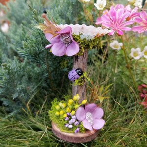 Op de afbeelding: Een miniatuur fee&euml;ntuin sc&egrave;ne met een paarse bloem, een lieveheersbeestje en een kleine houten basis met mos en gele bloemen.