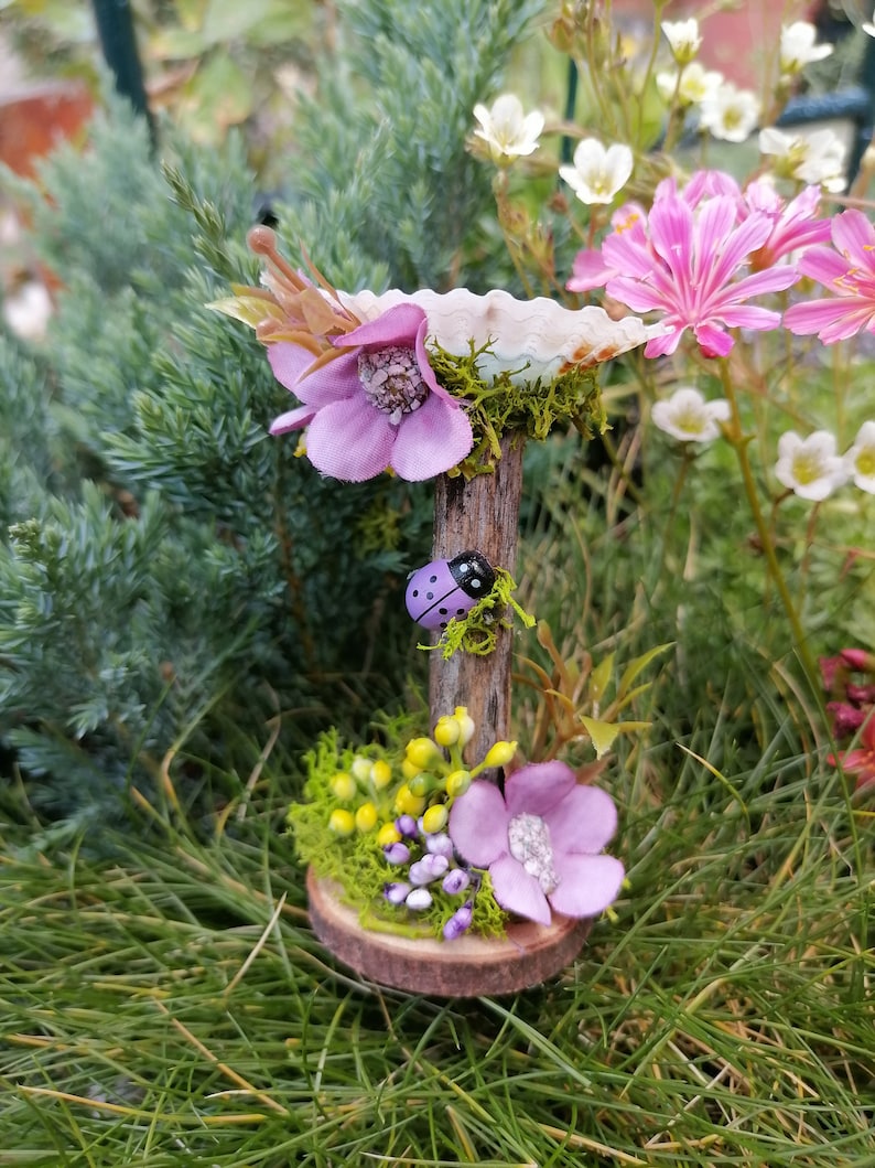 Op de afbeelding: Een miniatuur fee&euml;ntuin sc&egrave;ne met een paarse bloem, een lieveheersbeestje en een kleine houten basis met mos en gele bloemen.