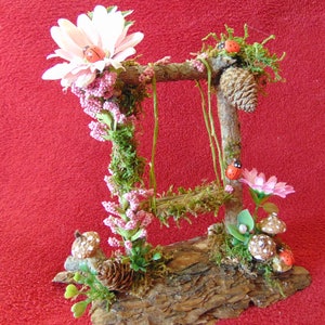 May include: A miniature fairy garden swing set made from twigs and moss, decorated with pink flowers, ladybugs, and mushrooms. The swing set is set on a piece of bark.