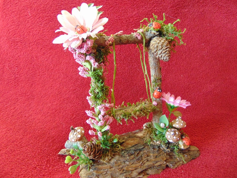 May include: A miniature fairy garden swing set made from twigs and moss, decorated with pink flowers, ladybugs, and mushrooms. The swing set is set on a piece of bark.