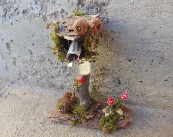 Può includere: Una cassetta delle lettere in miniatura realizzata con corteccia e rametti con un pezzo di carta arrotolato all'interno. La cassetta delle lettere è decorata con muschio, ghiande e una coccinella. Ci sono due piccoli funghi rossi con puntini bianchi alla base della cassetta delle lettere.