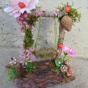 May include: A miniature fairy garden scene with a wooden swing set, pink flowers, green moss, and red ladybugs. The scene is set on a brown log base.