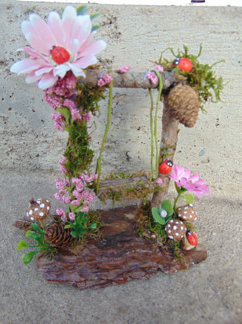 May include: A miniature fairy garden scene with a wooden swing set, pink flowers, green moss, and red ladybugs. The scene is set on a brown log base.