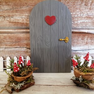 May include: Two miniature fairy doors made of wood with a red heart and a gold handle. The doors are decorated with a wooden grain pattern. The doors are placed in front of a wooden background with a white picket fence. Each door is decorated with a small potted plant with red and white mushrooms.