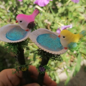 May include: Two miniature bird figurines perched on twig-like sticks with blue glitter-filled seashell bird baths. The pink bird is on the left and the yellow bird is on the right.