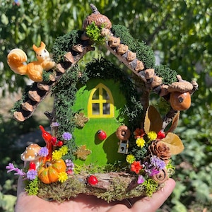 May include: A miniature fairy house with a green door and yellow window. The house is decorated with a squirrel, pumpkins, mushrooms, and colourful flowers. The roof is made of twigs and moss, creating a charming woodland scene.