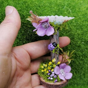 Op de afbeelding: Een miniatuur fee&euml;ntuin sc&egrave;ne met een paarse bloem, een lieveheersbeestje en een kleine houten basis met mos en gele bloemen.