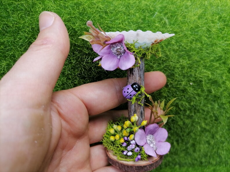 Op de afbeelding: Een miniatuur fee&euml;ntuin sc&egrave;ne met een paarse bloem, een lieveheersbeestje en een kleine houten basis met mos en gele bloemen.