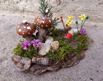 Può includere: Una scena di giardino fatato in miniatura con due funghi marroni con macchie bianche, circondati da muschio verde, piccole rocce e fiori colorati. Una minuscola coccinella riposa su una piccola base di legno, aggiungendo un tocco stravagante.