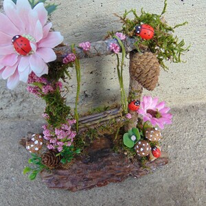 May include: A miniature fairy garden swing set made from twigs and moss, with pink flowers, ladybugs, and a pine cone.