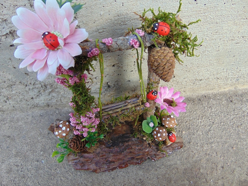 May include: A miniature fairy garden swing set made from twigs and moss, with pink flowers, ladybugs, and a pine cone.