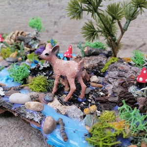 May include: A miniature woodland diorama featuring a small fawn figurine standing near a blue stream. The scene includes miniature trees, red and white spotted mushrooms, and various types of moss and lichen, all arranged on a piece of wood.
