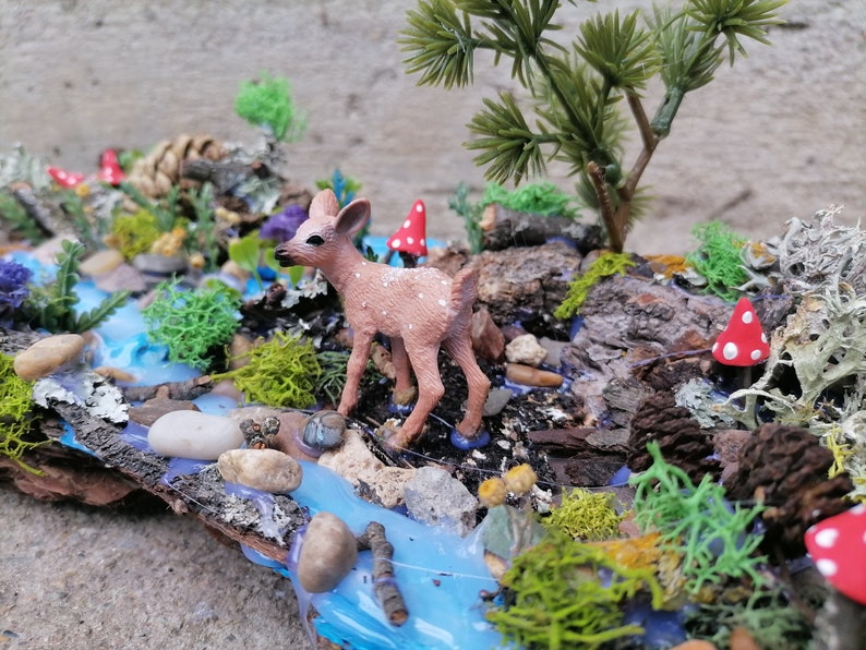 May include: A miniature woodland diorama featuring a small fawn figurine standing near a blue stream. The scene includes miniature trees, red and white spotted mushrooms, and various types of moss and lichen, all arranged on a piece of wood.