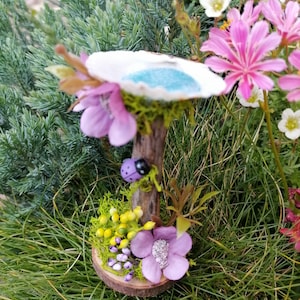 Op de afbeelding: Een miniatuur fee&euml;ntuin sc&egrave;ne met een klein blauw vijvertje, een paarse bloem en een lieveheersbeestje op een houten tak. De sc&egrave;ne is omgeven door groen gras en bloemen.