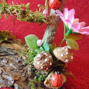 May include: A miniature woodland scene with a brown log base, green moss, pink flowers, and white-dotted mushrooms. Two red ladybugs are perched on the branches.