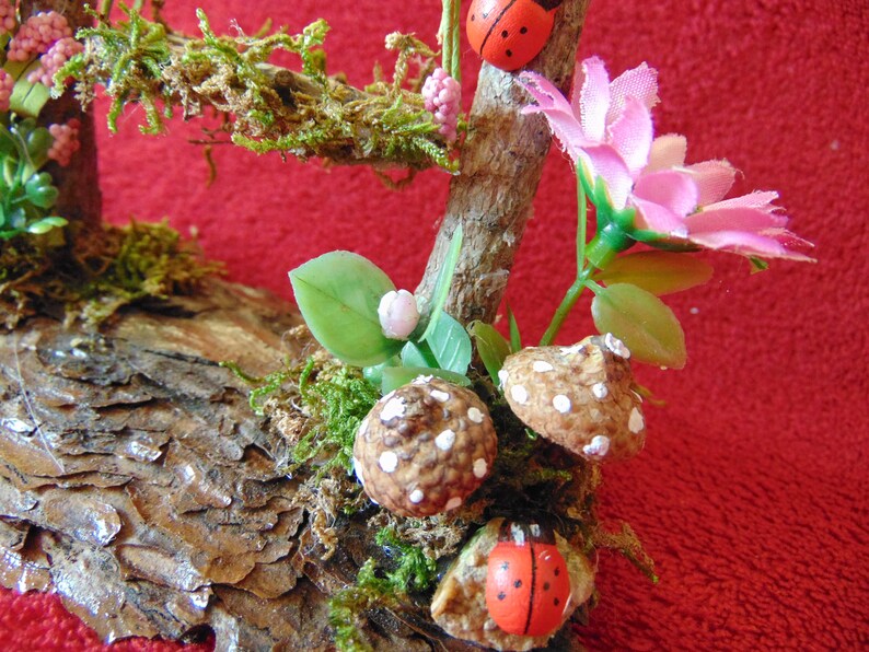 May include: A miniature woodland scene with a brown log base, green moss, pink flowers, and white-dotted mushrooms. Two red ladybugs are perched on the branches.