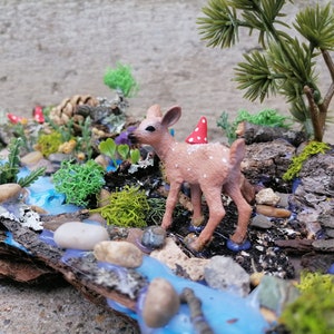 May include: A miniature woodland scene featuring a small fawn with a red and white mushroom hat, standing near a blue stream. The scene includes miniature trees, rocks, moss, and flowers, all arranged on a piece of wood.