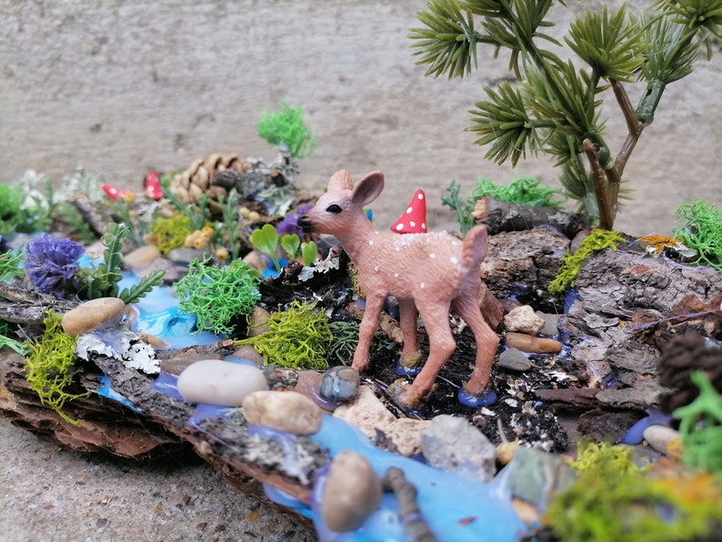 May include: A miniature woodland scene featuring a small fawn with a red and white mushroom hat, standing near a blue stream. The scene includes miniature trees, rocks, moss, and flowers, all arranged on a piece of wood.
